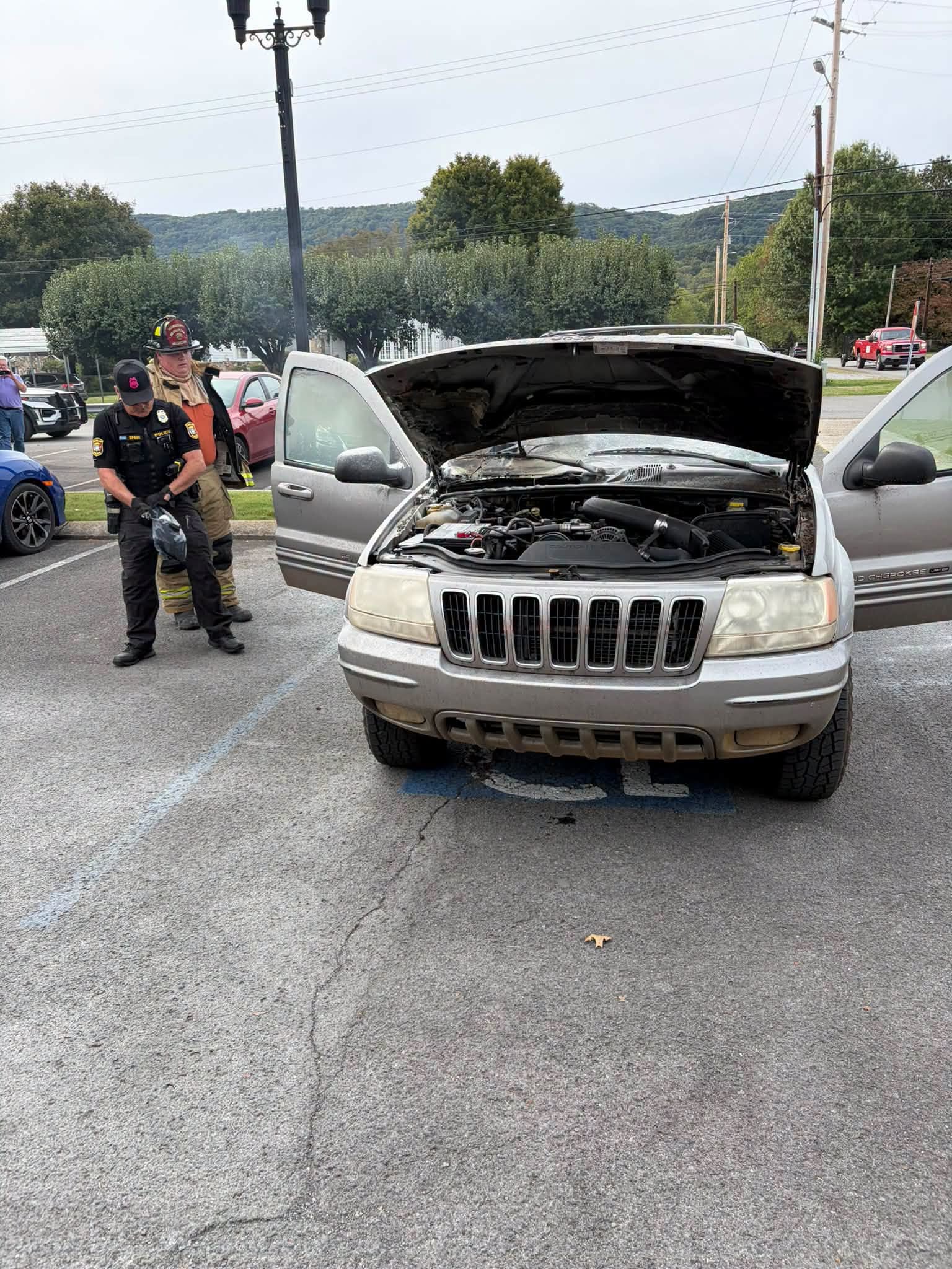 Dunlap: Jeep Cherokee gris con el cofre abierto en un estacionamiento tras el incendio, con un policía y un bombero al fondo